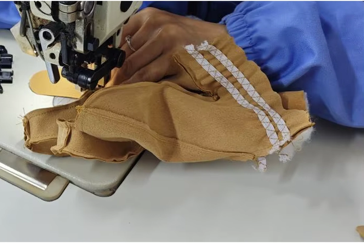 Hands guiding tan glove panels beneath a sewing-machine needle